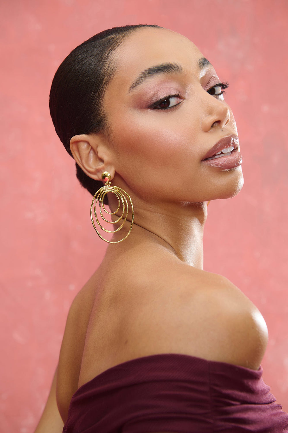 Pendientes dorados Nox de Miphai con diseño de aros concéntricos de acabado martillado, lucidos por una modelo con recogido y hombros descubiertos sobre fondo rosa.