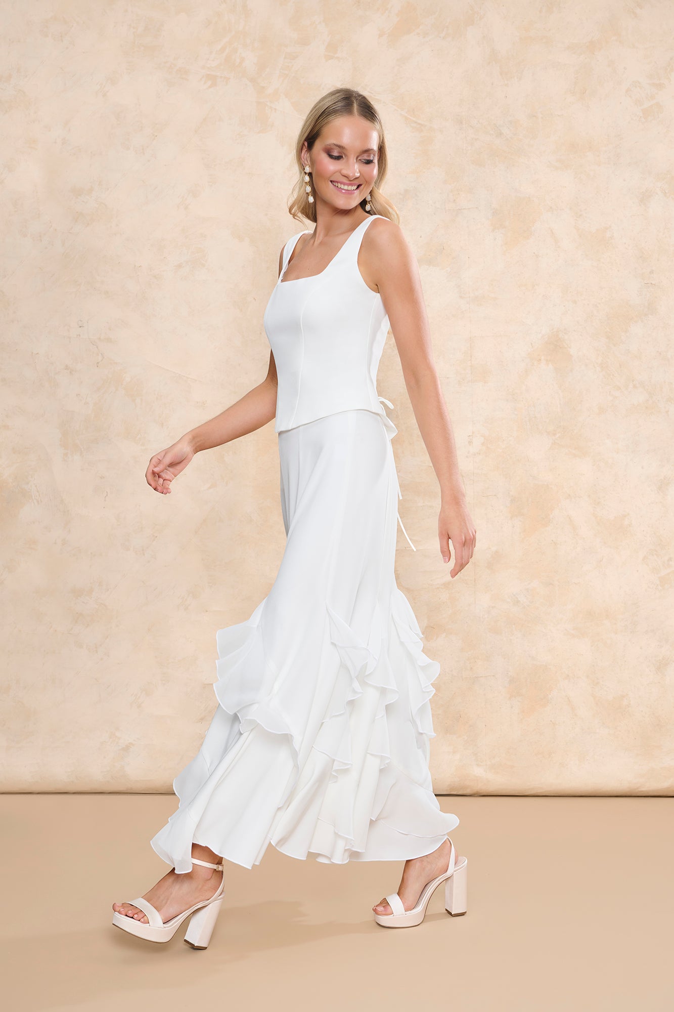 Fotografía de frente del conjunto "Samara" de Miphai en color blanco. El look consta de una falda larga de silueta fluida que destaca por sus llamativos volantes verticales en cascada desde la rodilla. Completan el conjunto unas sandalias de plataforma blancas y pendientes largos, sobre un fondo neutro.