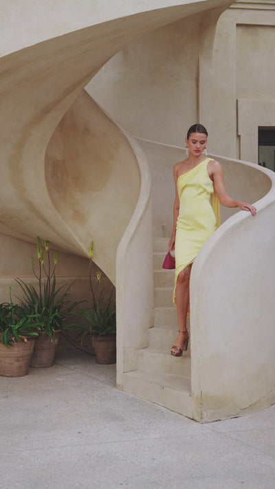 Vestido largo Lotus en crepé satén amarillo, con escote asimétrico, capa y espalda con tiras cruzadas.